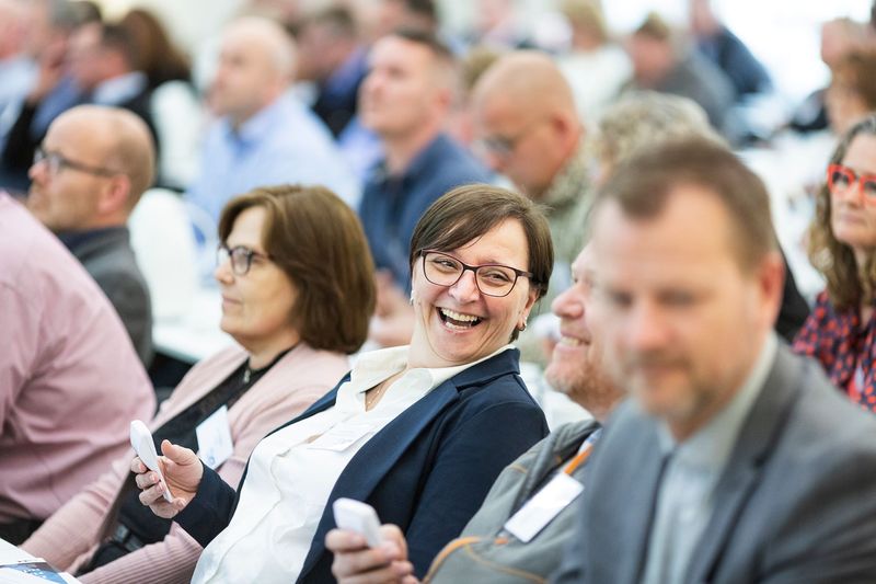 A woman in a conference audience turns to her neighbour, laughing.