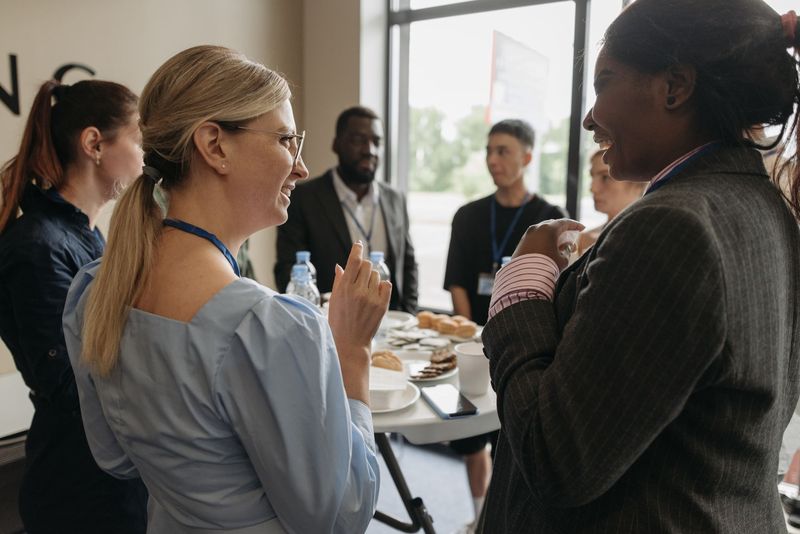 Two women wearing lanyards laugh together at a coffee break while other attendees talk around a table behind them.