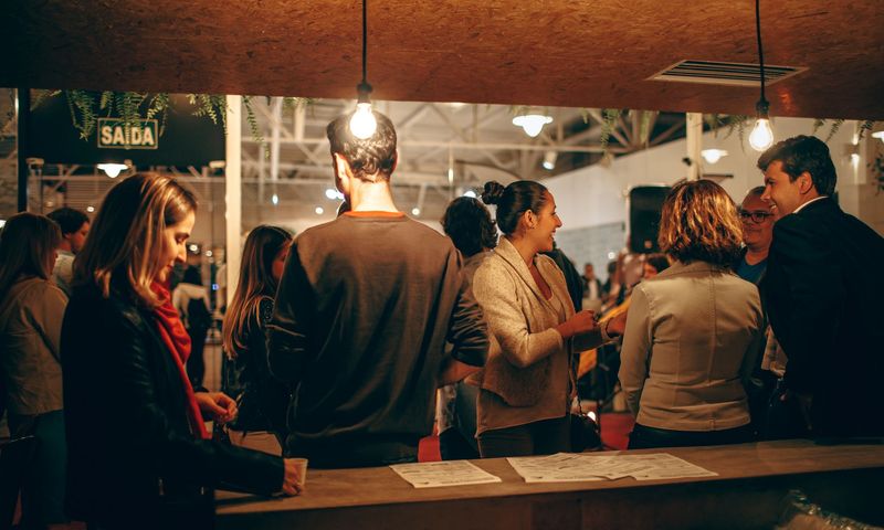 People chat at a counter under bare bulbs in a busy exhibition hall.