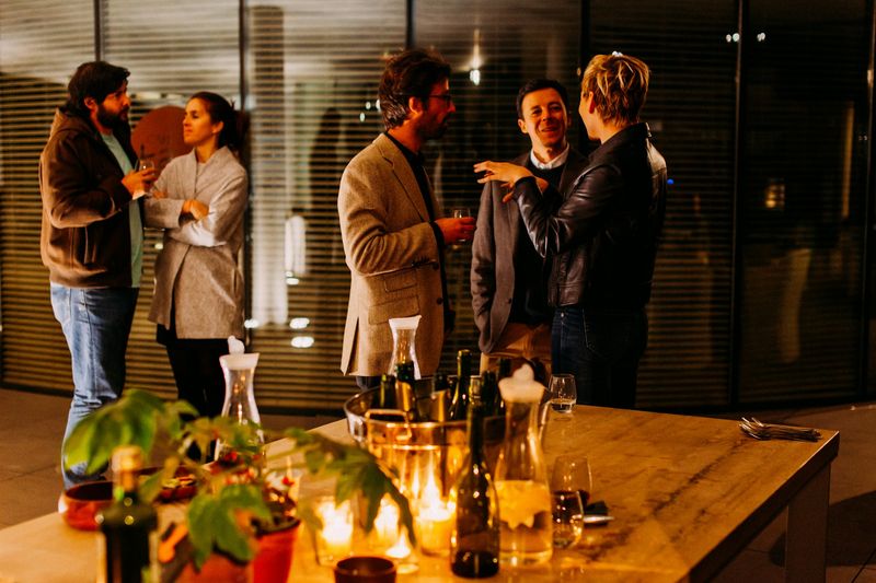Five people with drinks talk in two small groups beside a candle-lit table at an evening event.
