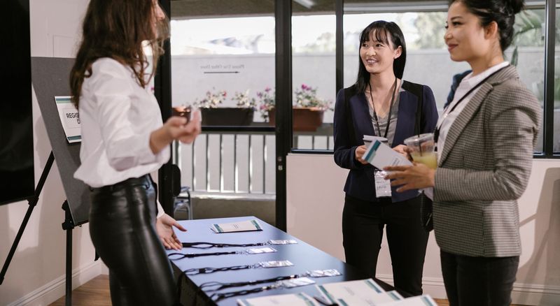 Three women talk across a registration desk laid out with lanyards and programmes.
