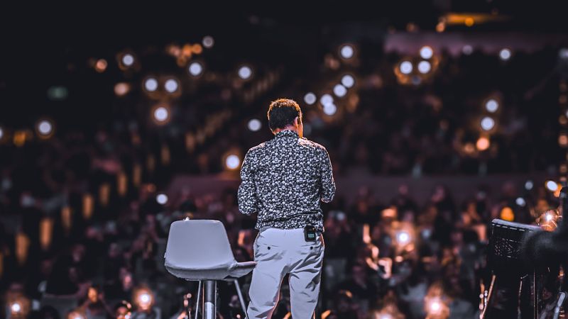 A speaker seen from behind on stage, facing a large audience holding up phone lights.